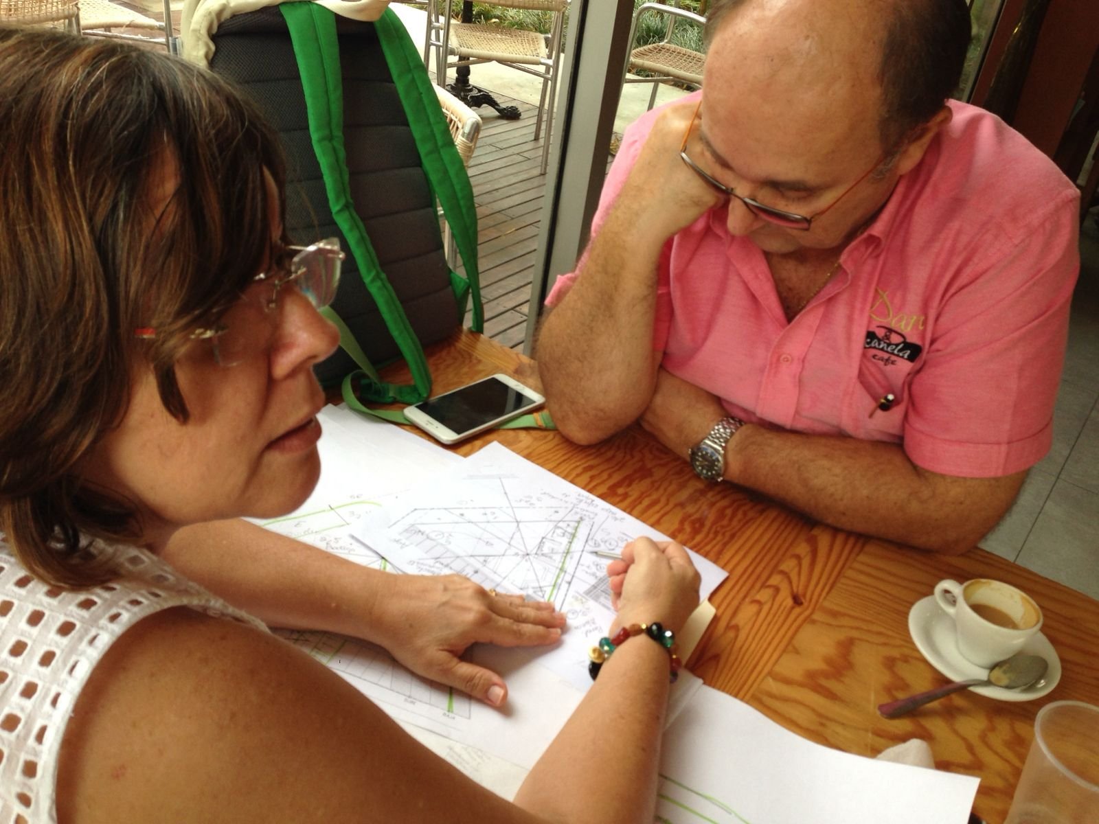 A woman and a man sitting at a wooden table looking at architectural plans, with a coffee cup beside them.