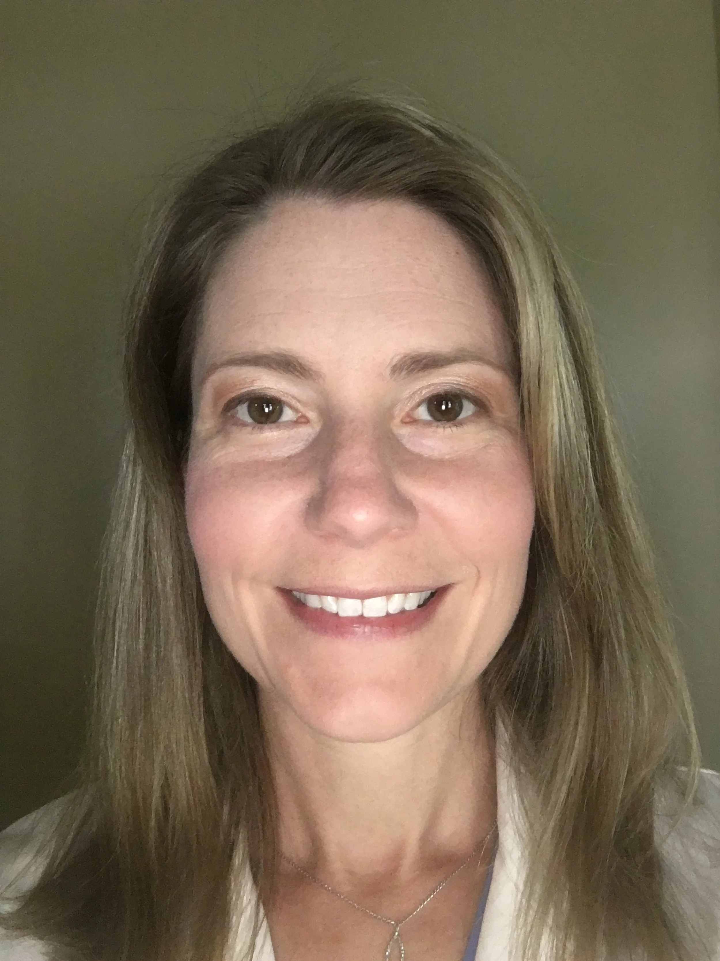 A woman with shoulder-length light brown hair and brown eyes smiling at the camera, wearing a light-colored top and a silver necklace, with a plain background.
