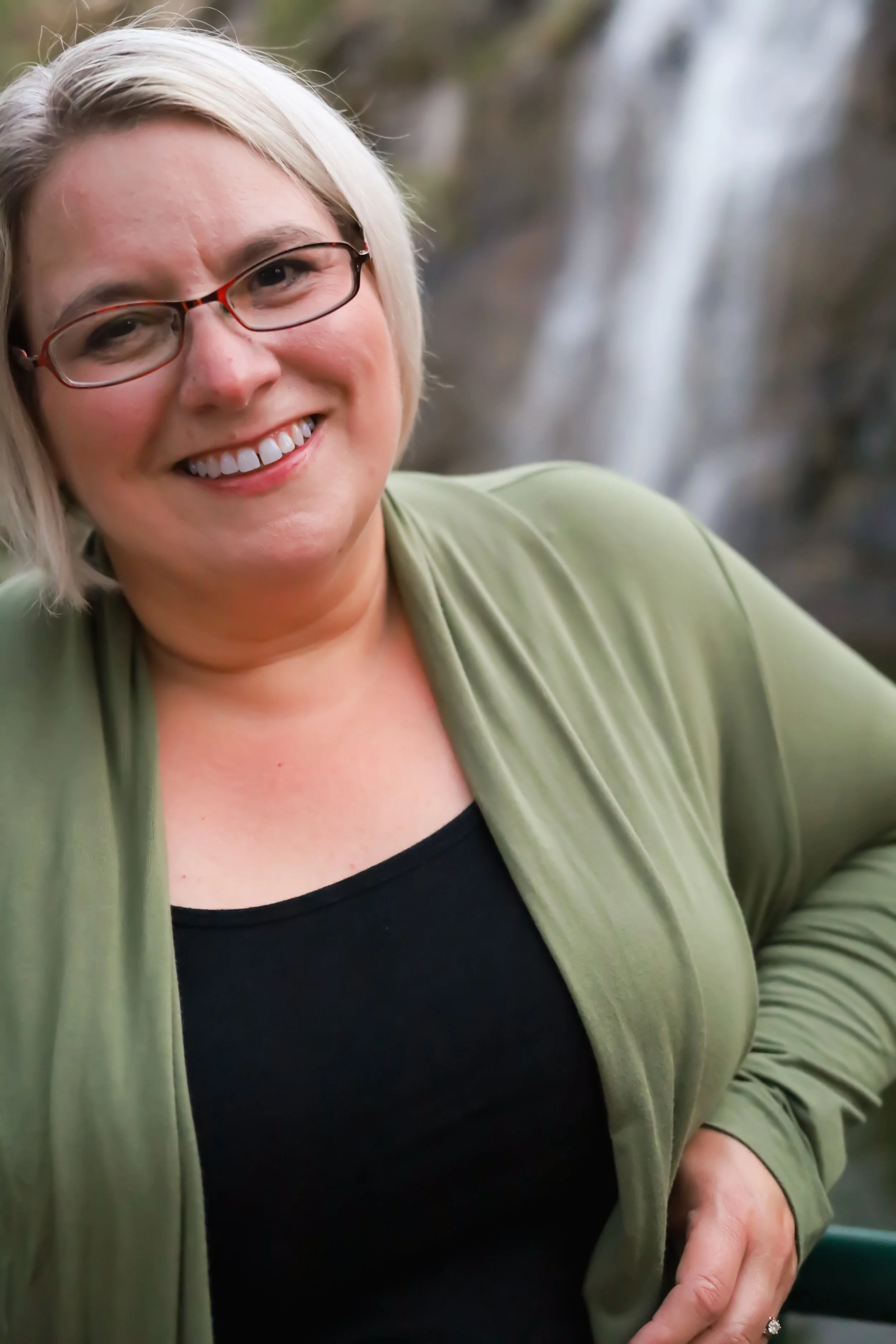 A smiling woman with blonde hair and glasses, wearing a black top and green jacket, standing outdoors near a waterfall.