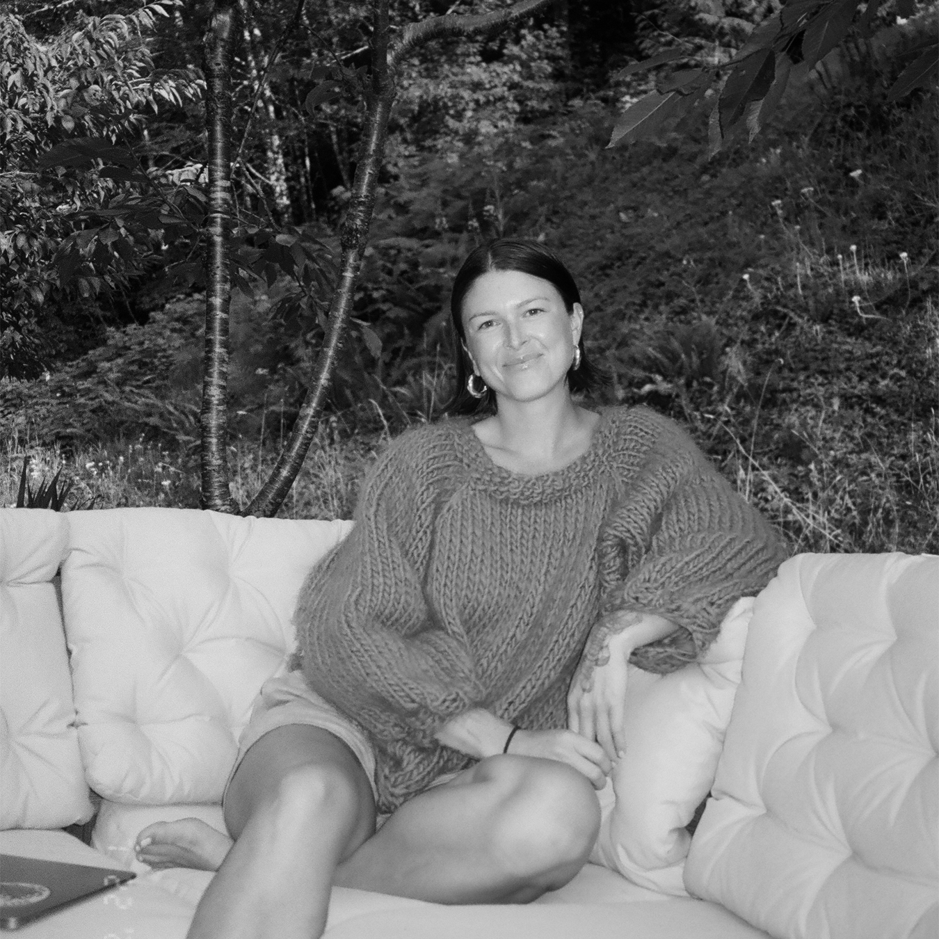 A woman with dark hair smiling while sitting on a cushioned outdoor sofa with her legs crossed, wearing a chunky knit sweater. She is surrounded by a lush, wooded background.
