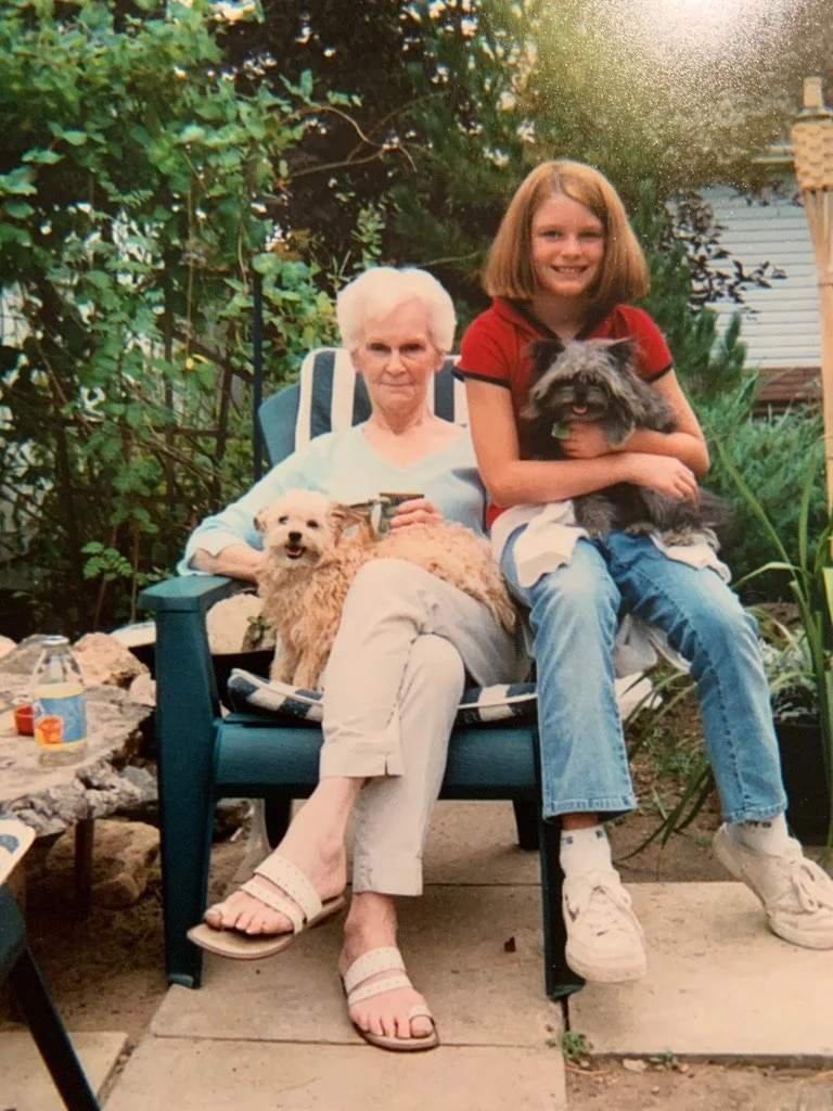 An elderly woman and a young girl sitting outdoors on a blue chair, each holding a small dog, with lush greenery in the background.