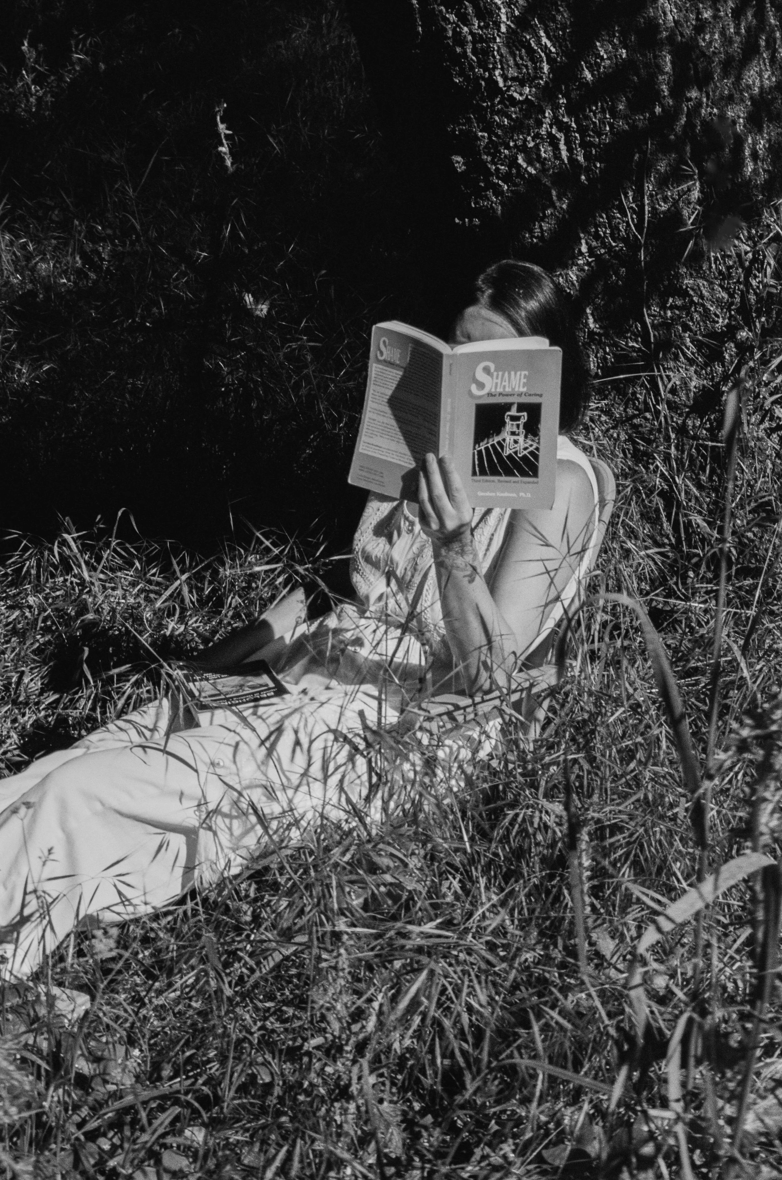 A person sitting on the ground in tall grass, holding a magazine titled 'Shane,' partially hiding their face, with trees and foliage in the background.