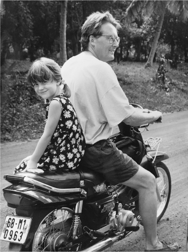 A black and white photo of a person and a young girl on a motorcycle riding on a dirt road, with trees in the background. The person is smiling, wearing glasses, a casual t-shirt, and shorts.