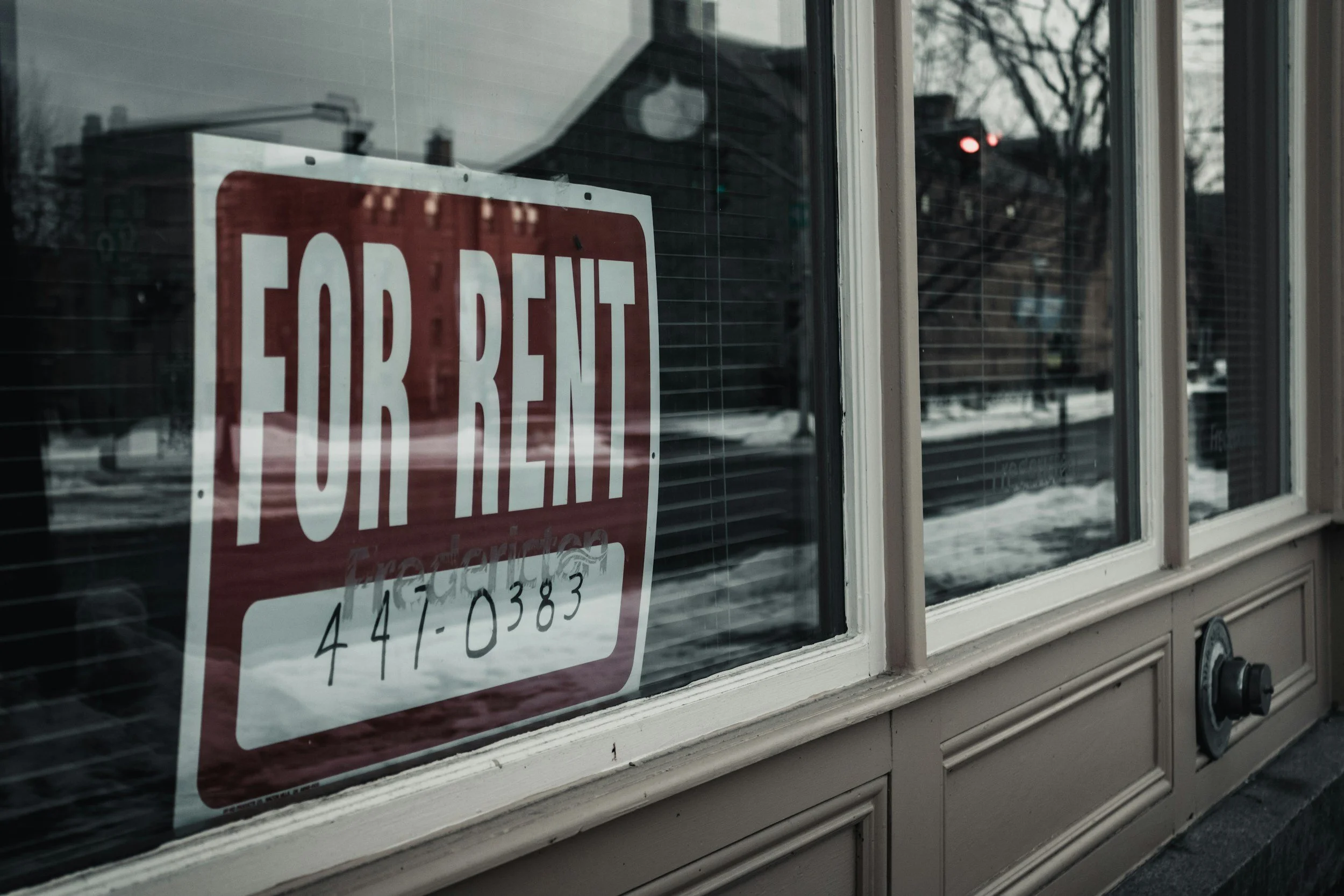 Close-up of a 'For Rent' sign posted on a window with blinds, reflecting the street and sky outside on a cloudy day.