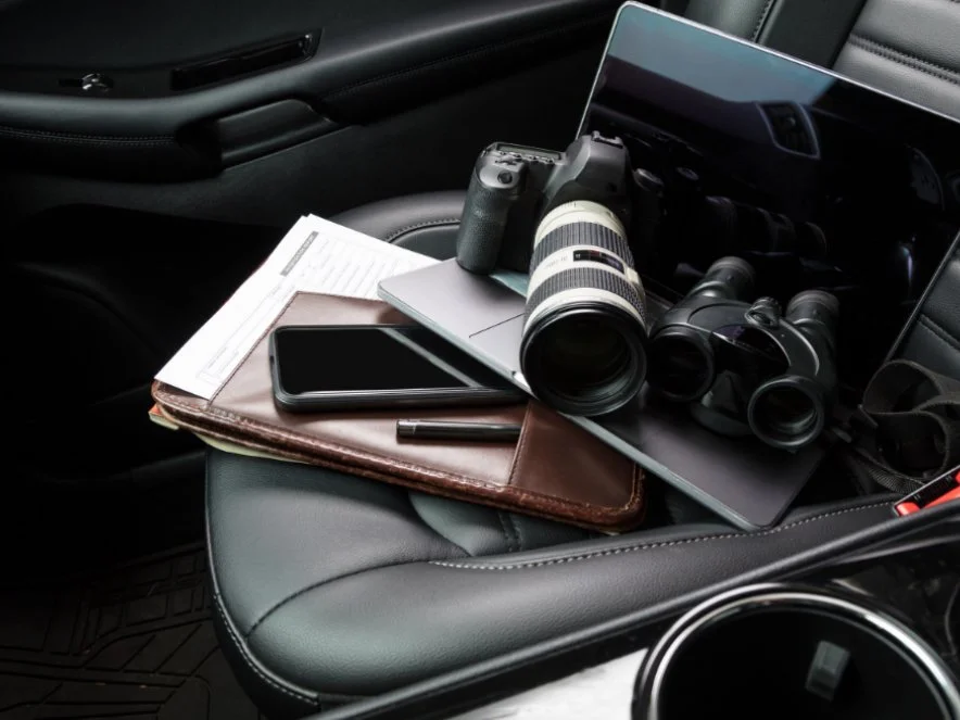 Camera with a telephoto lens, binoculars, a smartphone, a pen, paper, and a laptop on a car seat.