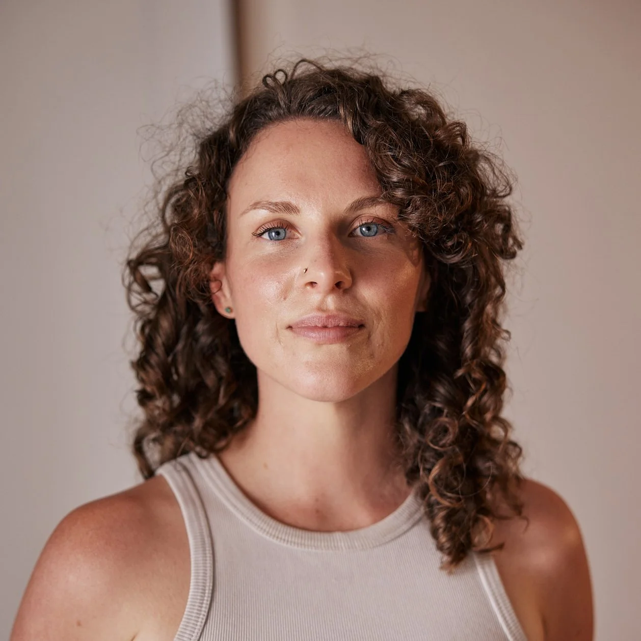 A woman with curly brown hair and blue eyes looking at the camera, wearing a light-colored sleeveless top, standing indoors against a neutral background.