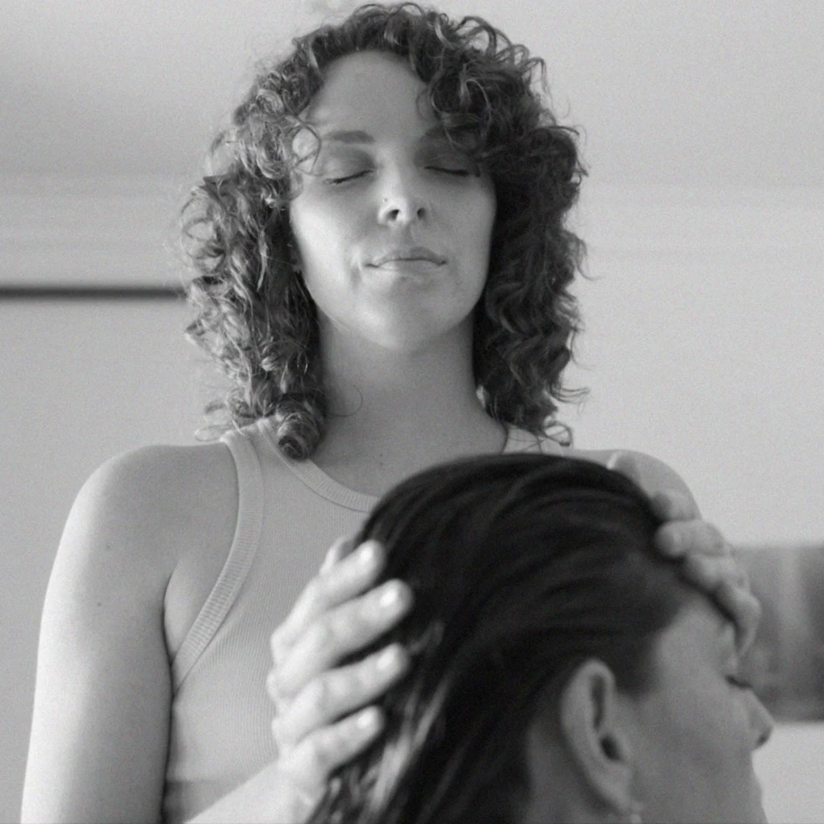 A woman with curly hair gently holding a person's head in a comforting gesture, both with closed eyes, in a calming and intimate moment.