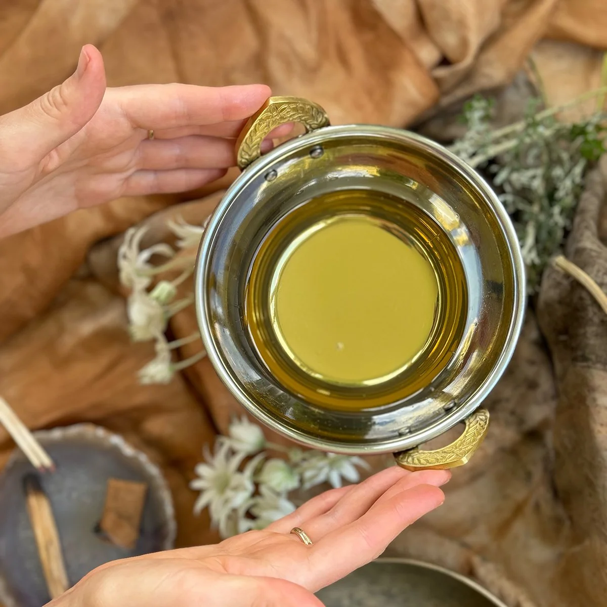A person holding a metallic cup filled with greenish herbal oil, with decorative gold handles, surrounded by dried herbs and flowers on a brown fabric surface.