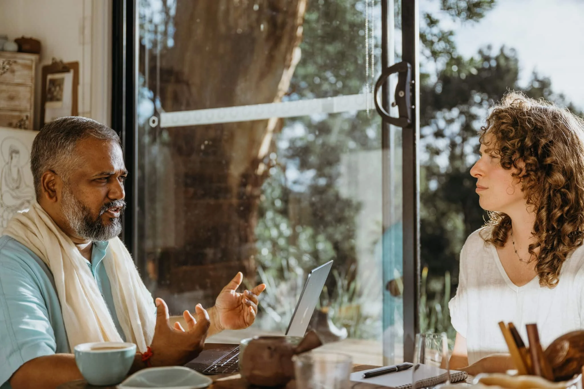 A man with gray hair and beard, wearing a light blue shirt and a white scarf, is speaking and gesturing with his hands while sitting at a table with a laptop, coffee cup, and some food. A woman with curly brown hair in a white shirt listens attentively. They are inside a room with large glass windows showing trees outside.