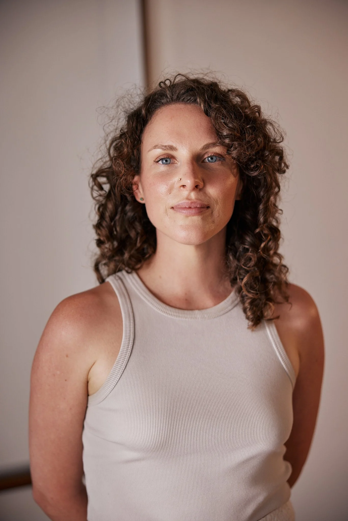 A young woman with curly brown hair and blue eyes, wearing a sleeveless beige top, standing indoors with a neutral expression.