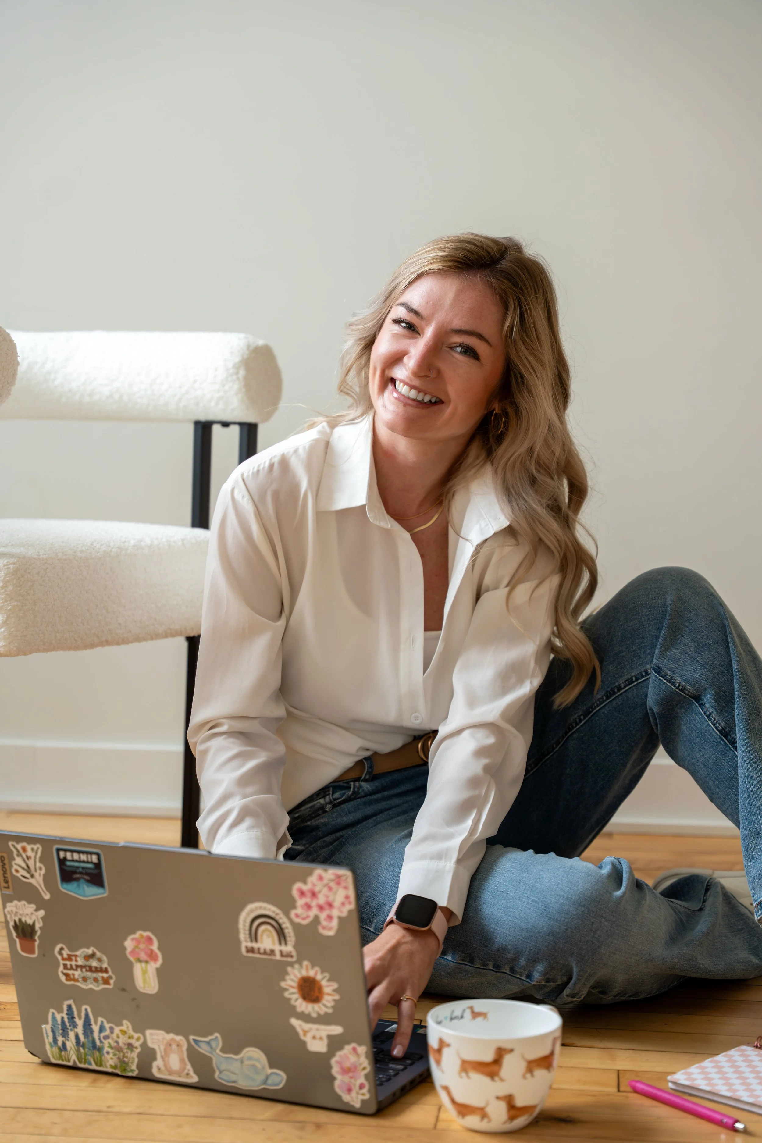 Smiling woman with blonde hair sitting on the floor using a laptop with colorful stickers, wearing a white shirt, jeans, and a smartwatch, in a room with wooden flooring and a white wall. There is a white cup with dog illustrations, a pink pen, and a notebook nearby.