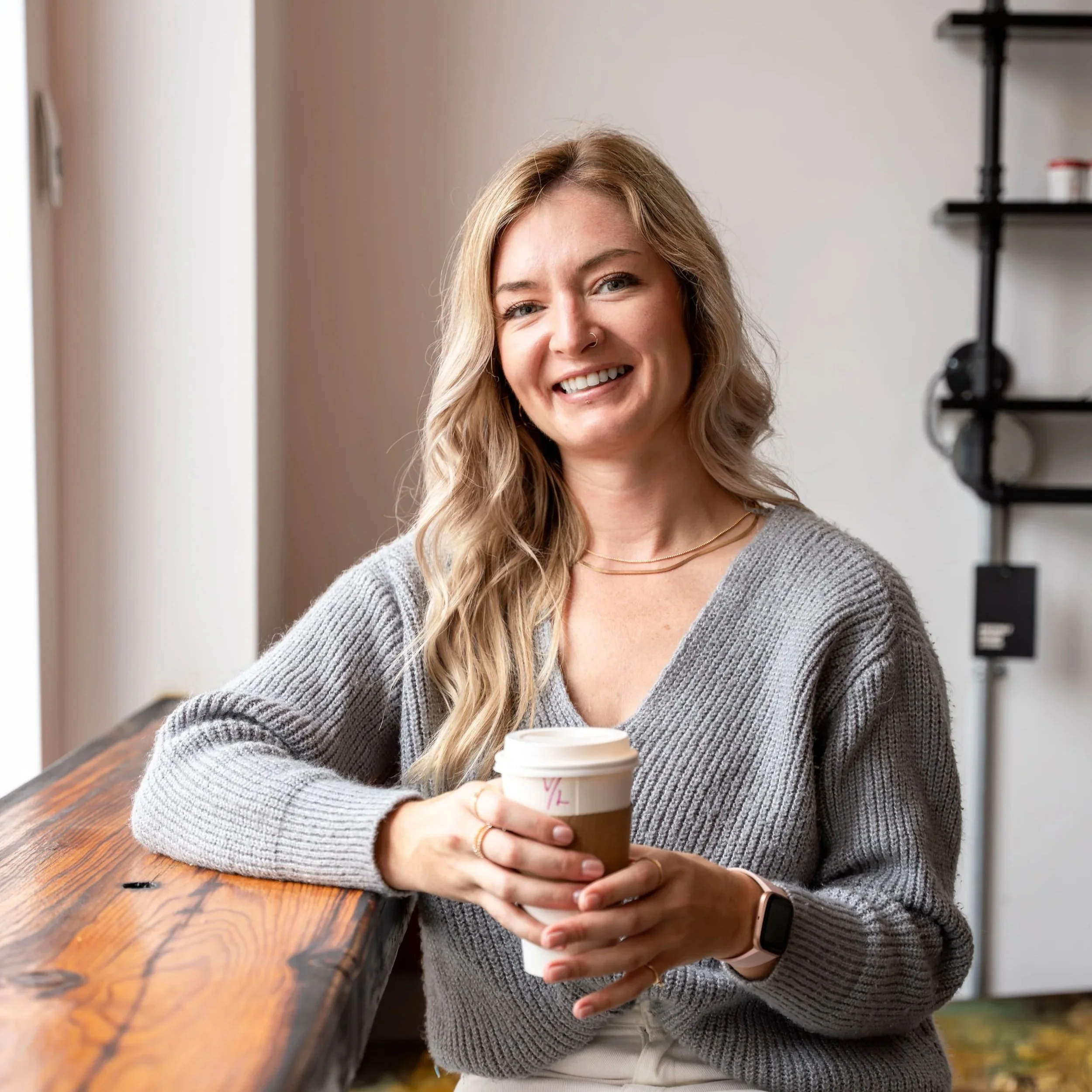 A woman with long wavy blonde hair sitting at a wooden table, smiling, holding a coffee cup in both hands, wearing a gray sweater and a smartwatch.