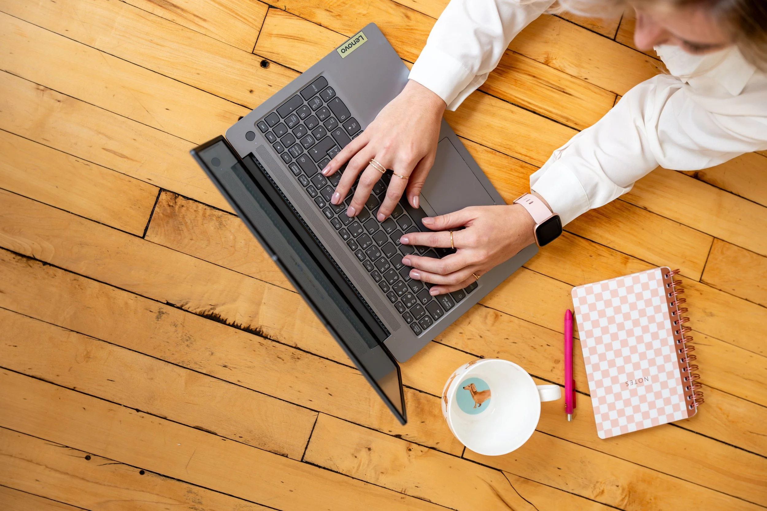 A person working on a laptop on a wooden floor with a pink checkered notebook, a pen, and a mug with a duck illustration.