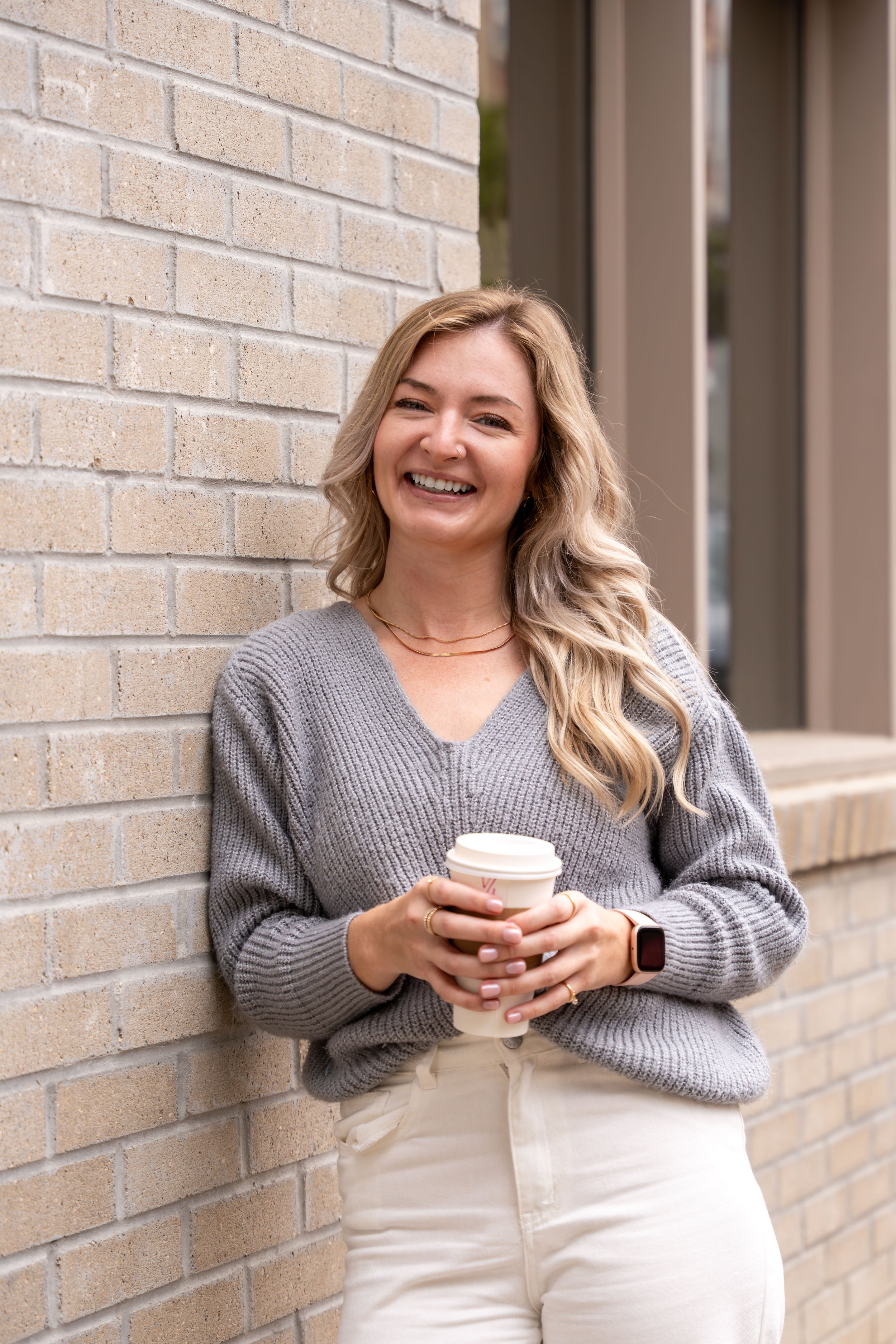 A young woman with wavy blonde hair smiling and holding a to-go coffee cup, leaning against a beige brick wall outside a building.