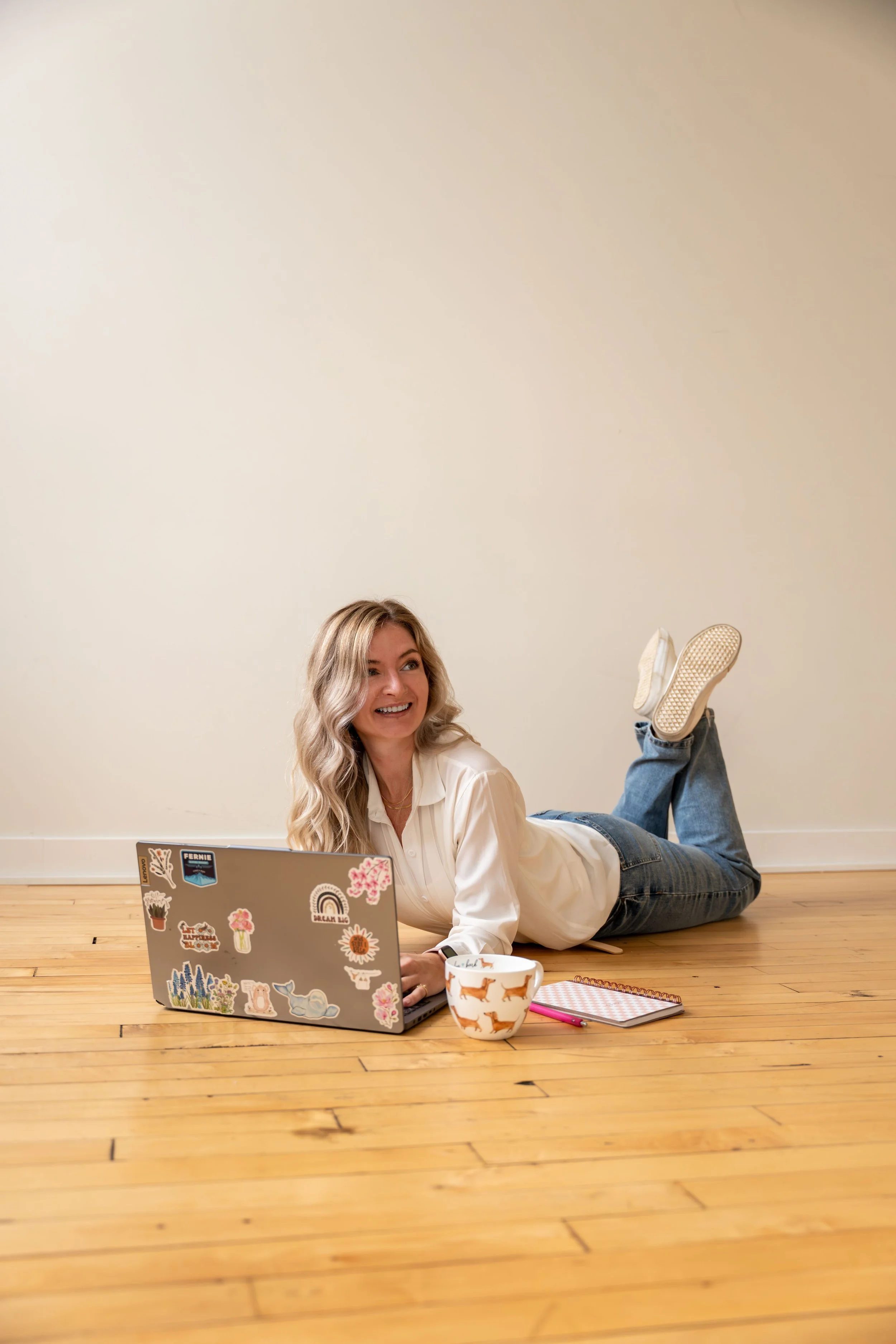 A woman with blonde hair smiling while lying on her stomach on a wooden floor, using a laptop with stickers, with a notepad, a pink pen, and a coffee mug nearby.