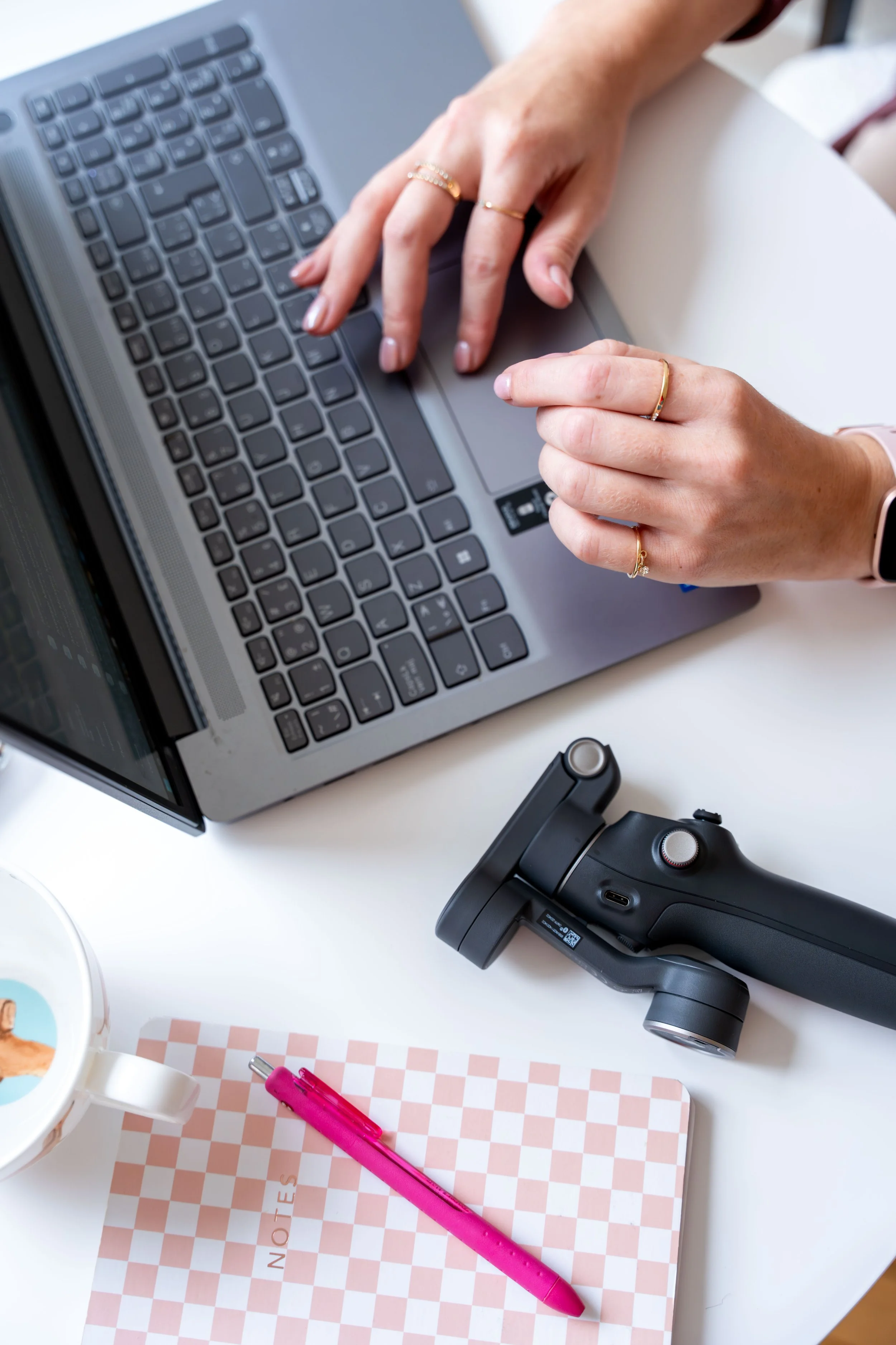 Close-up of a person’s hands typing on a gray laptop keyboard, with rings on fingers, a pink pen on a pink checkered notebook, a black massage gun, and a mug with a drawing on it on a white desk.