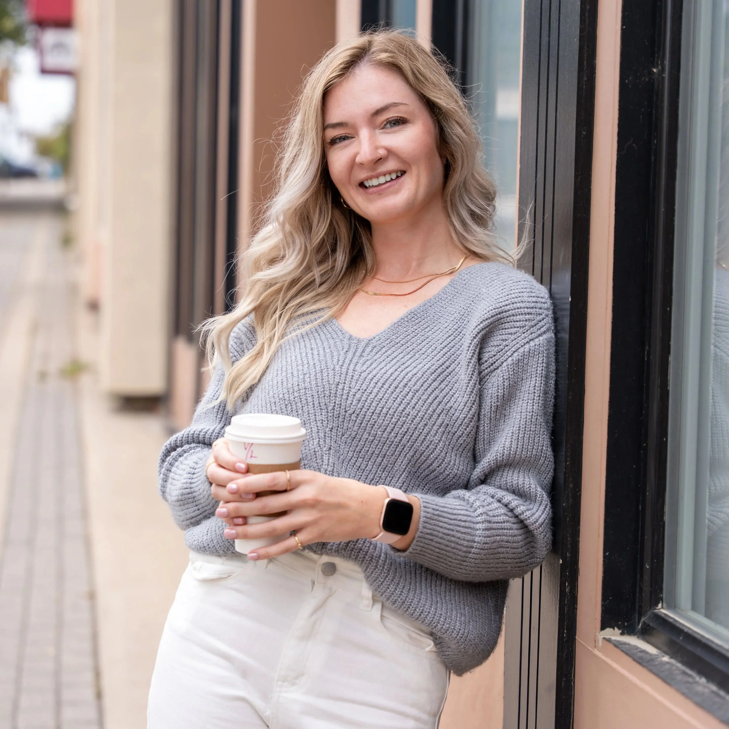 A young woman with long wavy blonde hair smiling outdoors, leaning against a window, holding a to-go coffee cup, wearing a gray knit sweater, white pants, a smartwatch, and jewelry.