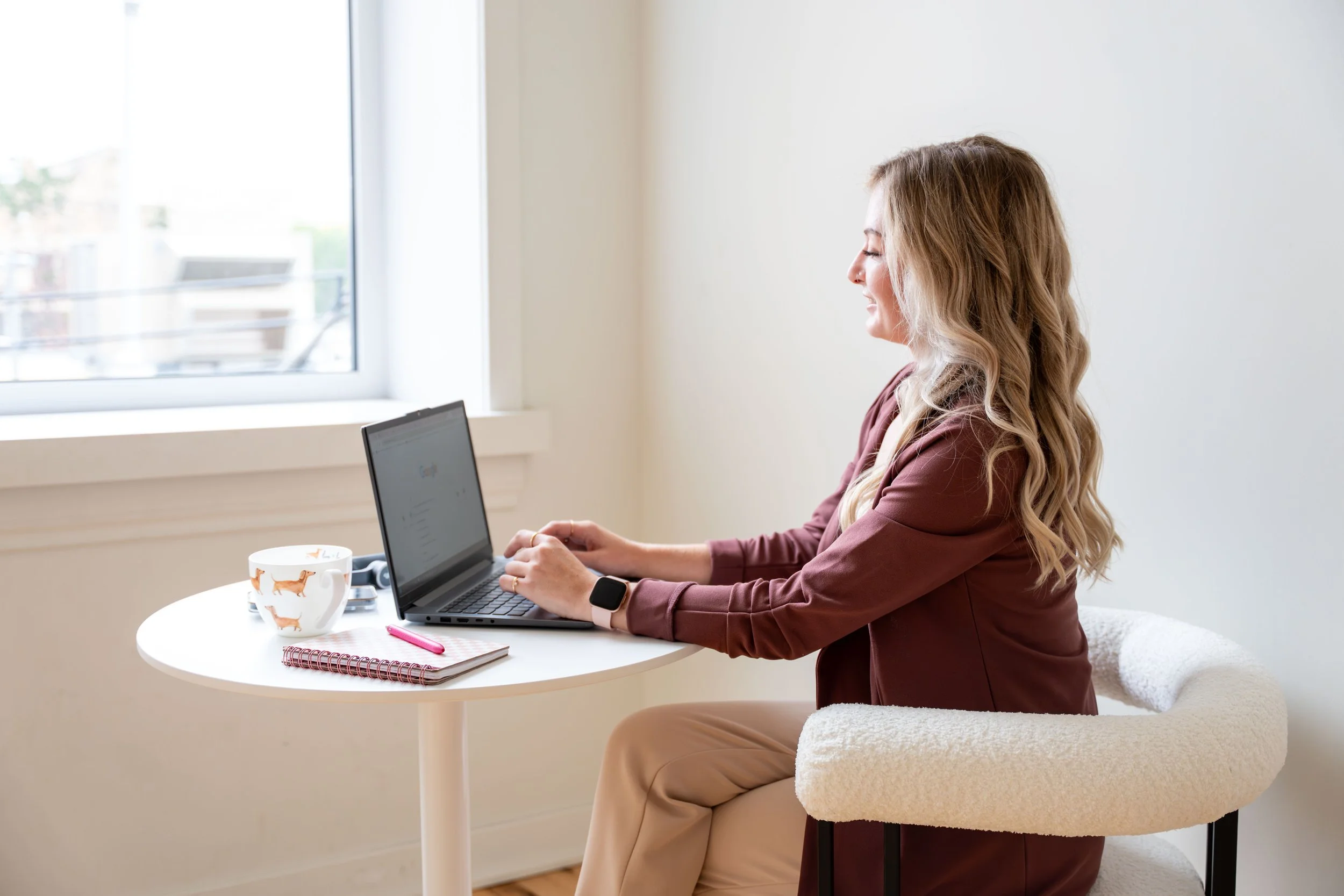 Woman smiling while working on a laptop at a white round table near a window on a sunny day.