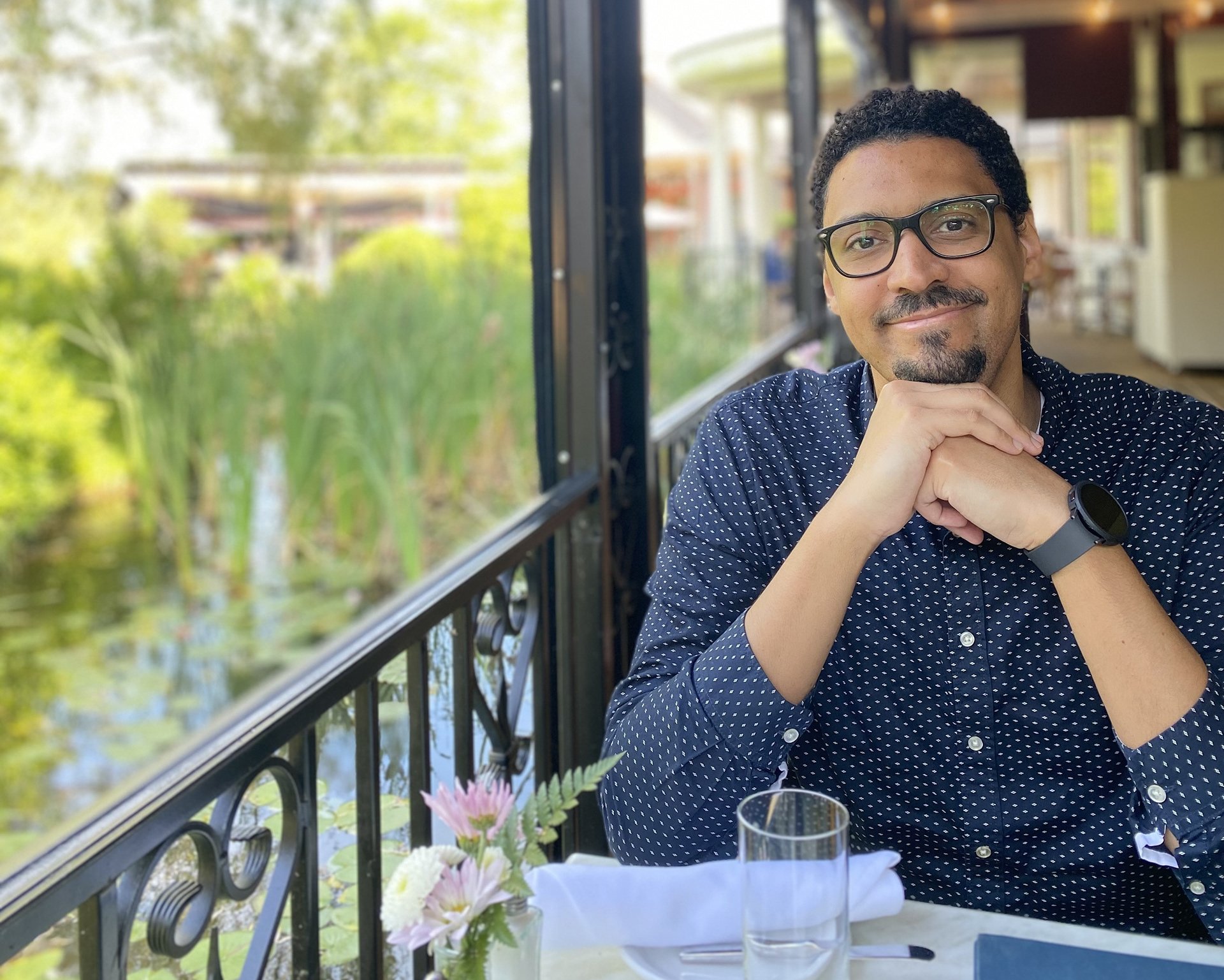 A man with glasses, black curly hair, sitting at a table with a view of greenery and a river outside. He is smiling, with his hands clasped under his chin, wearing a dark blue, polka-dotted shirt and a smartwatch.