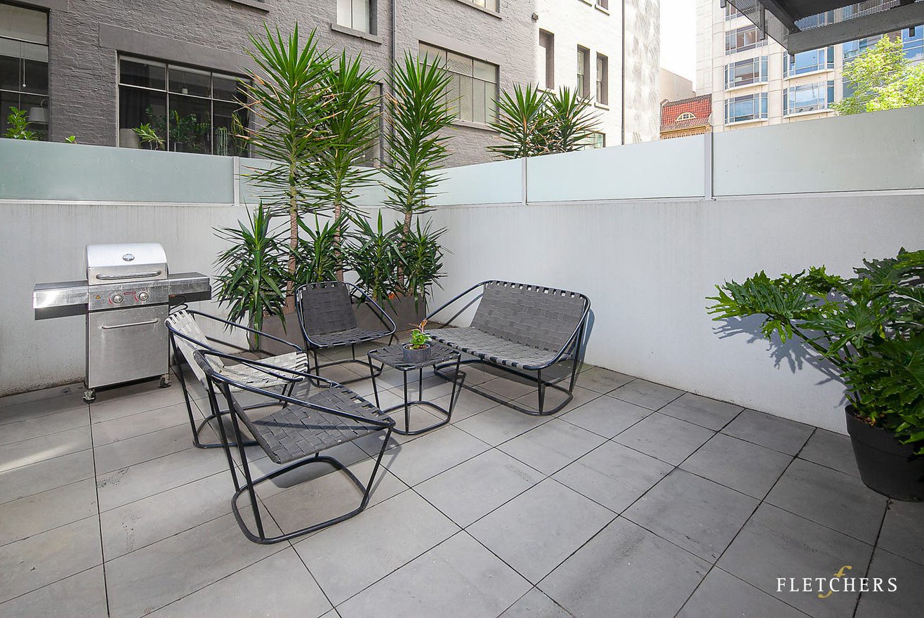 Outdoor patio with four black metal chairs, a small matching table, a grill, several tall green plants, and potted plants, enclosed by a white wall with city buildings in the background.