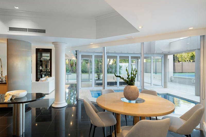 Modern living space with glass walls, circular dining table with six chairs, large potted plant, and pool outside