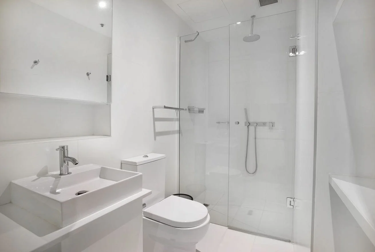 Modern white bathroom with a glass shower enclosure, a square vessel sink with a chrome faucet, a mirror, a toilet, and a small corner sink or bathtub on the right.
