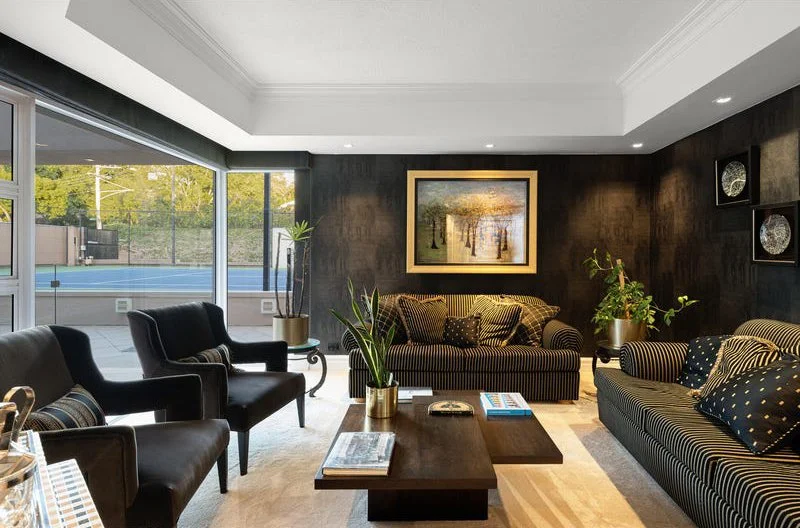 Living room with black and striped sofas, potted plants, artwork, and a view of a tennis court through large glass sliding doors.