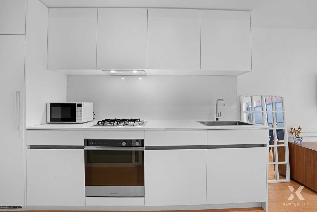 Minimalist white kitchen with microwave, stove, oven, and sink, with multiple mirrors leaning against the wall and potted plant on a wooden cabinet.