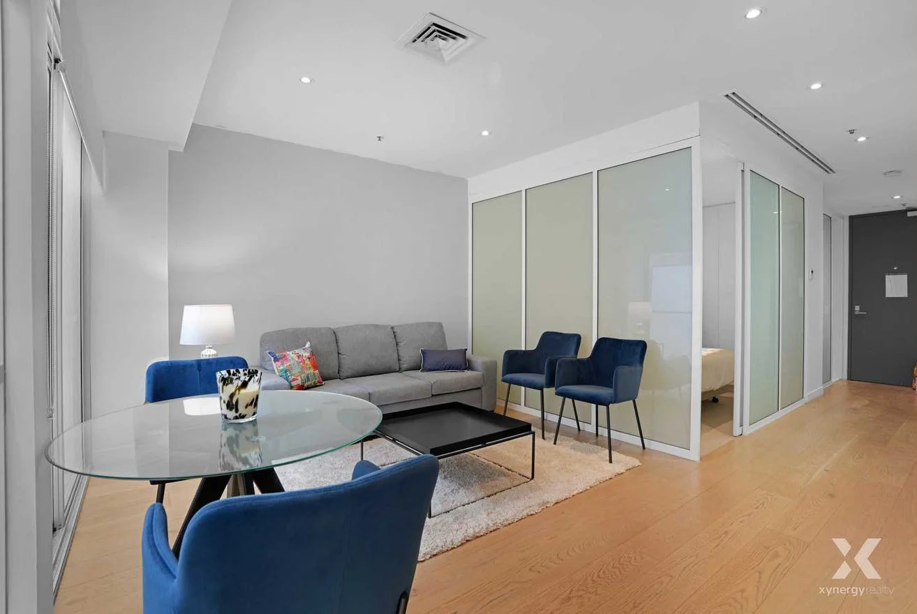 Modern living room with a gray couch, blue chairs, glass dining table, and wooden floors.