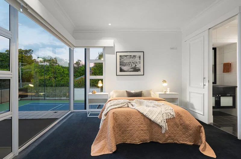 Bedroom with a sliding glass door leading to a patio and tennis court, white walls, bed with peach blanket and white throw, two bedside lamps, framed black and white artwork, view of trees outside.