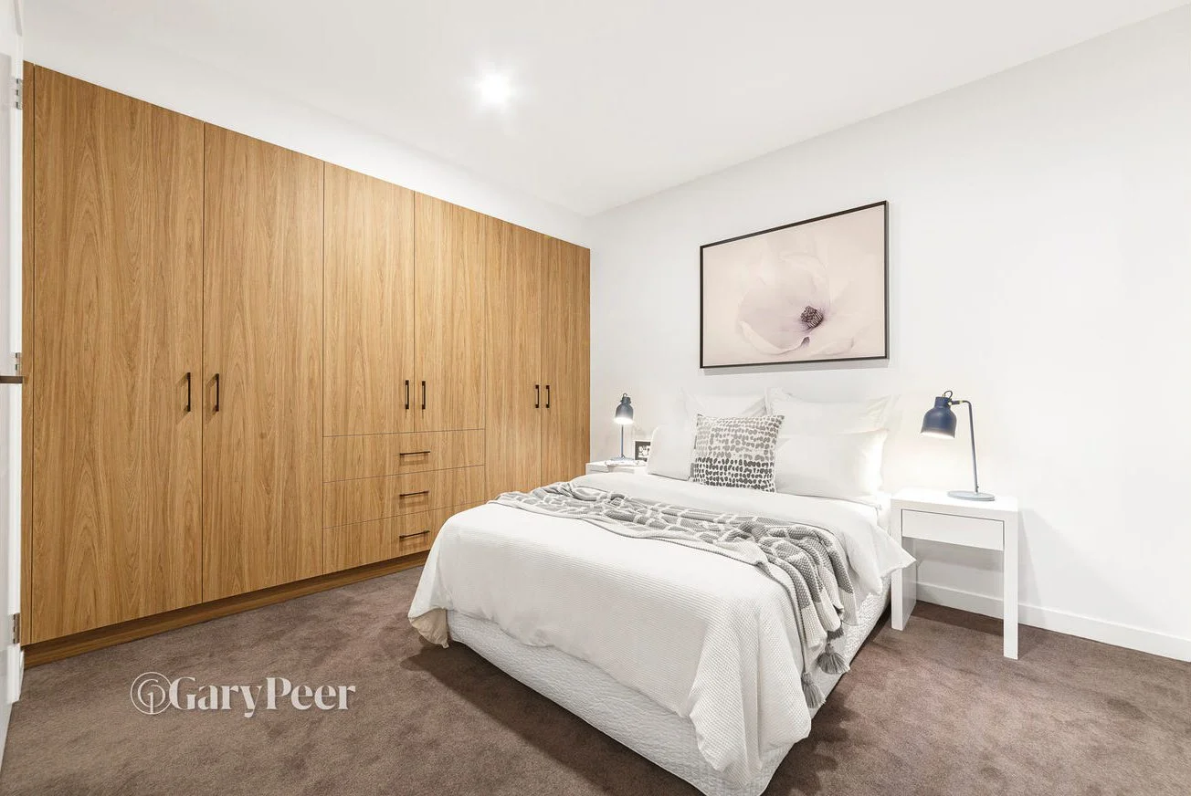 A modern bedroom with white walls, a white bed with gray and white patterned pillows, and a brown wooden wardrobe.