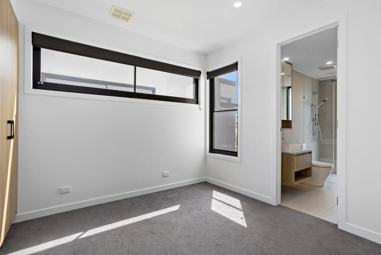 Empty bedroom with gray carpet, white walls, two black-framed windows, and a door leading to a bathroom with a glass shower, toilet, and wooden vanity.
