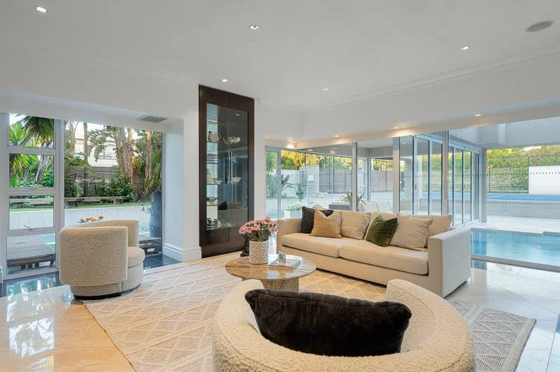 Modern living room with large windows, white walls, beige sofas, a round coffee table with flowers, and a view of an outdoor pool and garden.