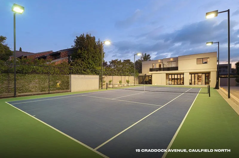 An empty tennis court at dusk inside a residential complex, with modern multi-story buildings in the background and tall lights illuminating the court.