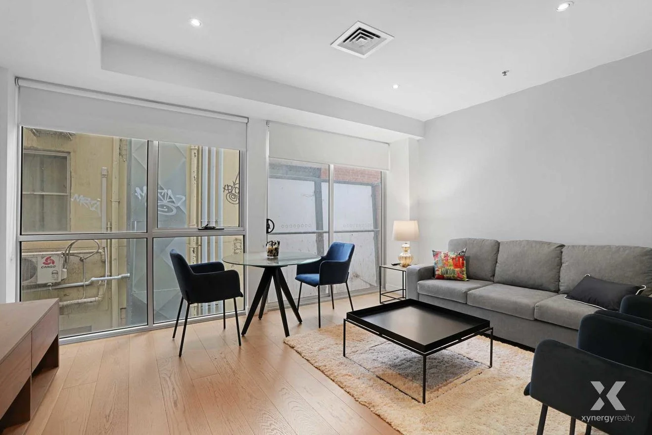 Modern living room with large windows, gray sofa, black coffee table, small round dining table, and two blue chairs, with a warm floor lamp and decorative cushions.