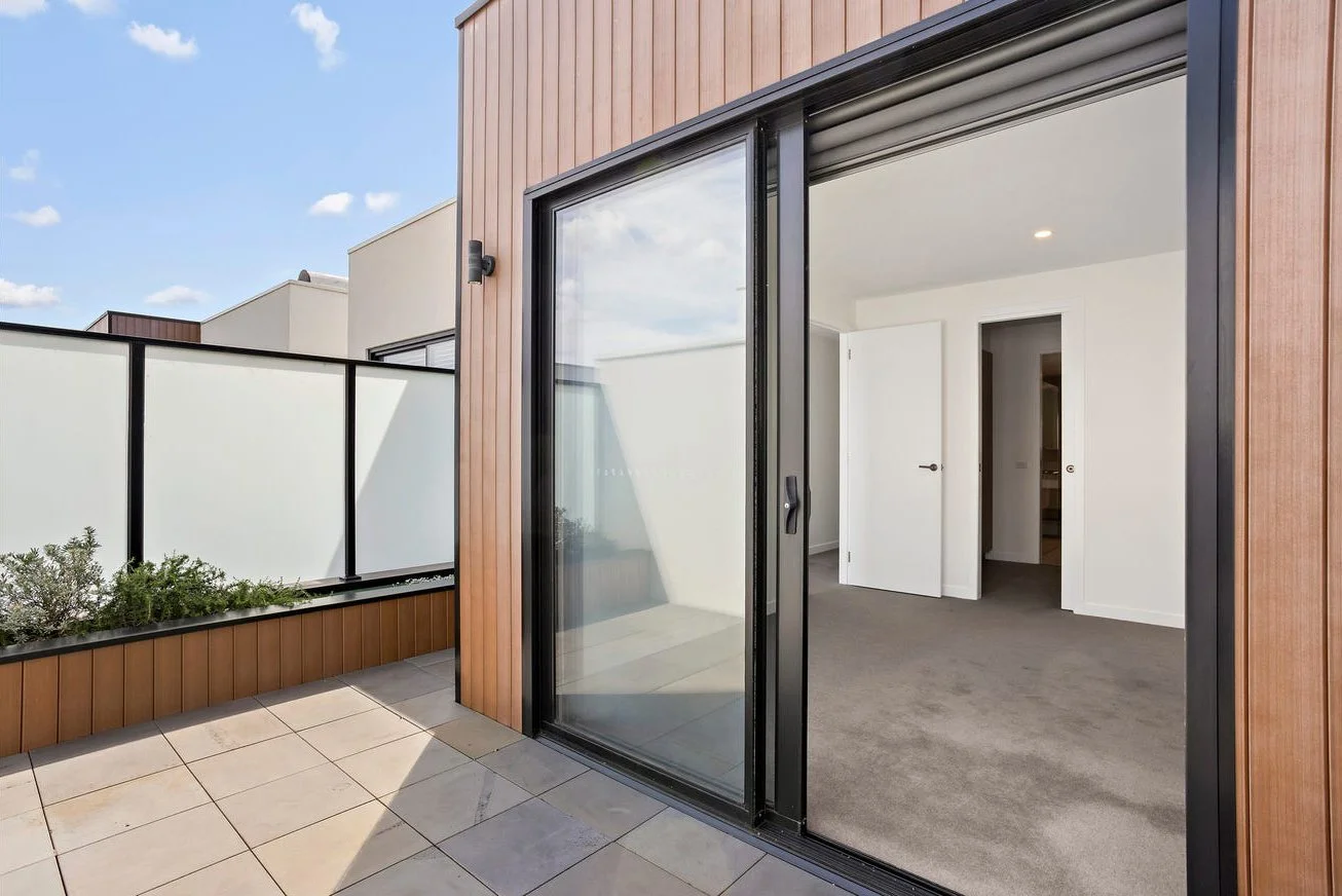 Modern apartment balcony with outdoor flooring, brown wood paneling, glass sliding door leading to a white-carpeted interior room, and a frosted glass fence with black framing.