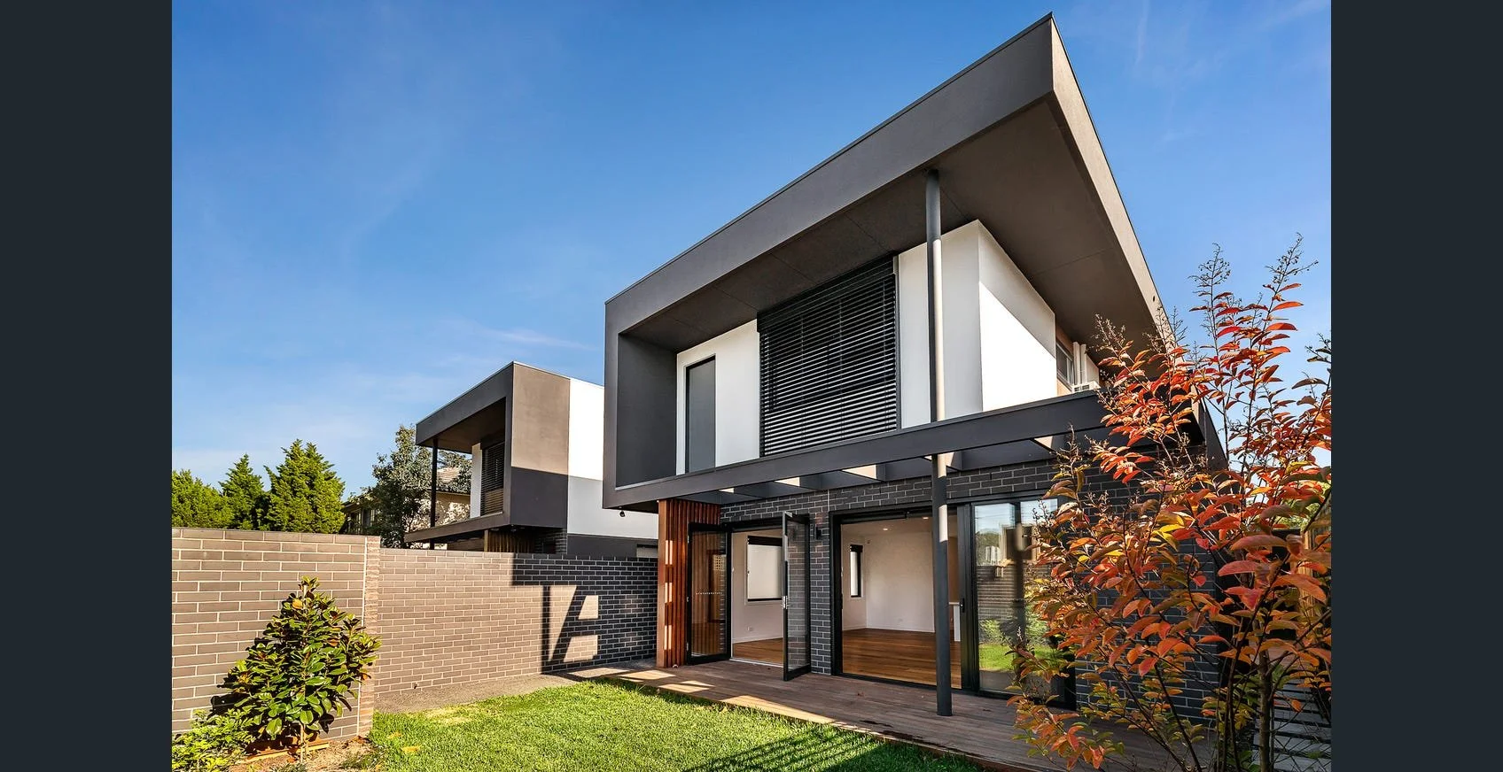 Modern two-story house with black brick and white exterior walls, large glass doors, and a small wooden deck, surrounded by a grassy yard and trees, under a clear blue sky.