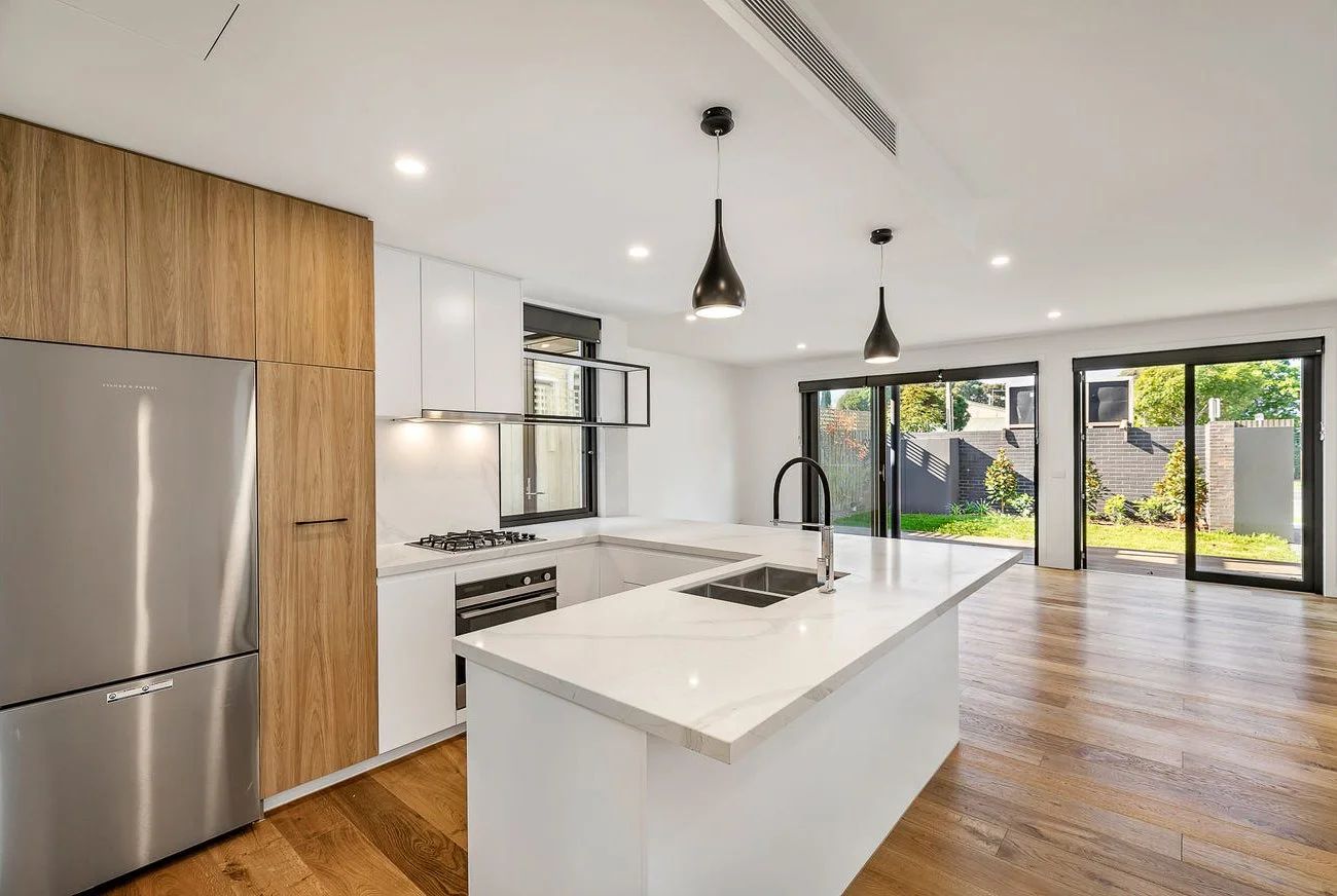 Modern kitchen with white countertops, wooden cabinets, stainless steel refrigerator, and black pendant lights, with a view of a backyard through large sliding glass doors.