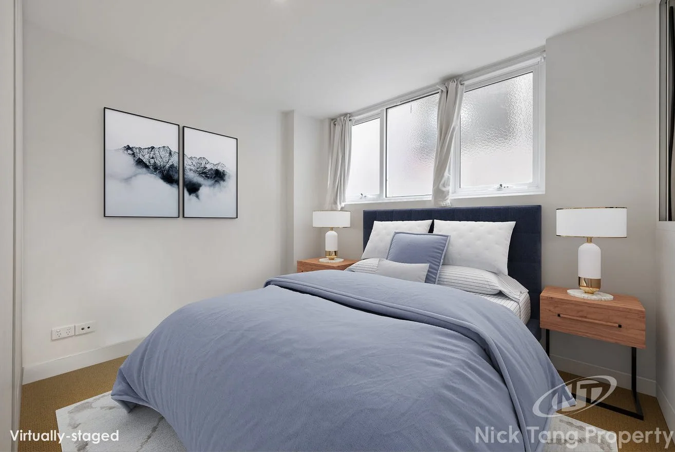 A neatly made bedroom with a bed, nightstands, lamps, and artwork, with windows allowing natural light.
