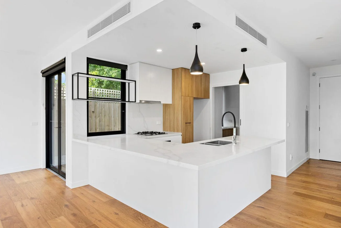 Modern kitchen with white countertops, black pendant lights, wooden cabinets, black-framed window, hardwood floors, and sliding glass door.
