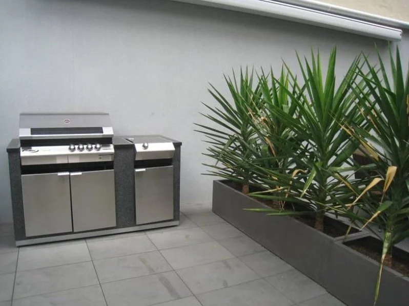 A stainless steel outdoor barbecue grill next to a large planter with green tropical plants.