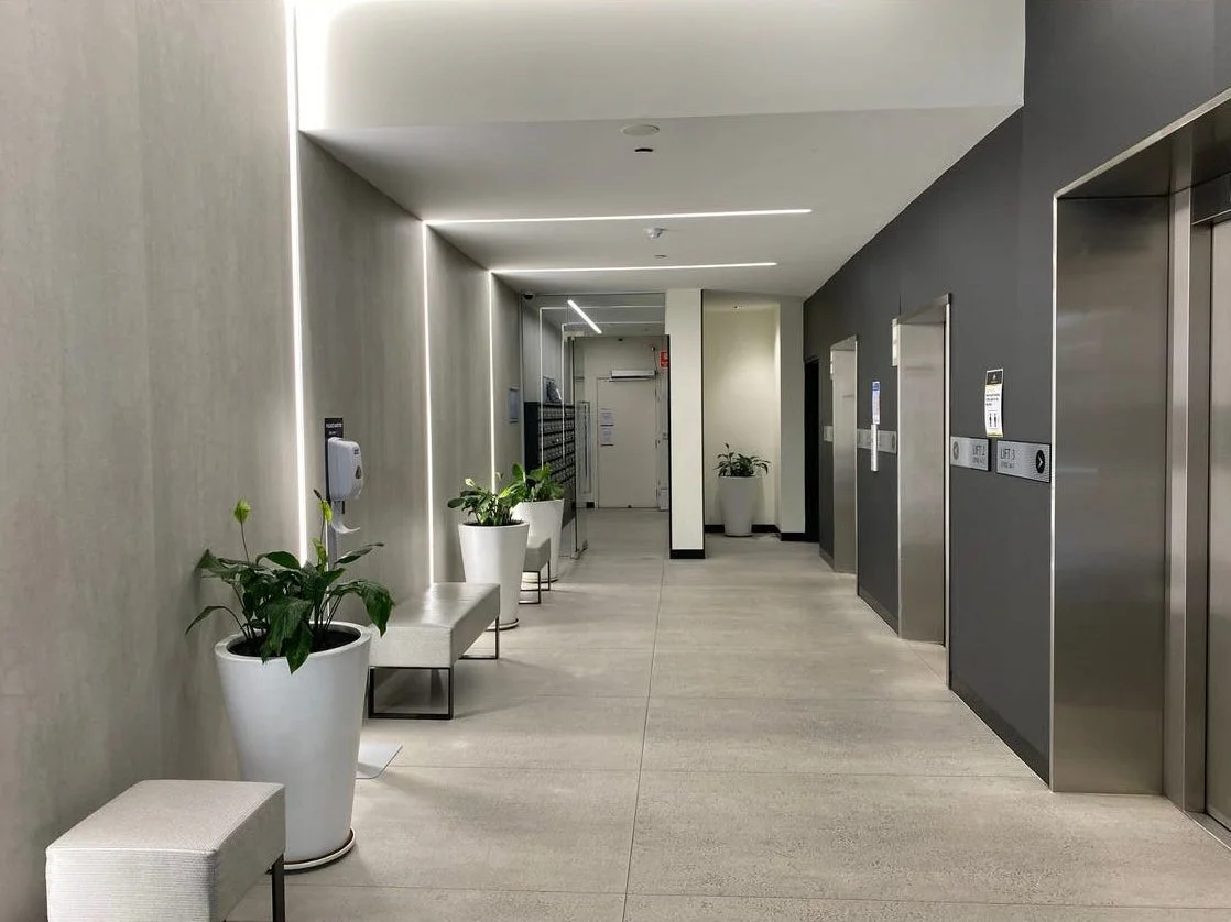 Modern office building lobby with elevators, potted plants, benches, and mailboxes.
