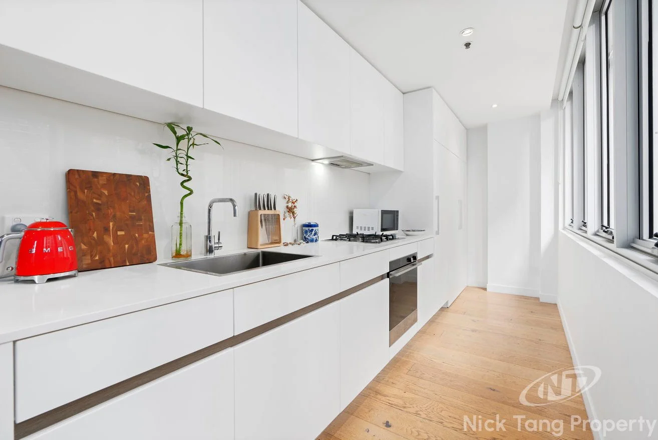 Modern, white kitchen with wooden floor, countertop, cabinets, oven, microwave, window, and various kitchen items.