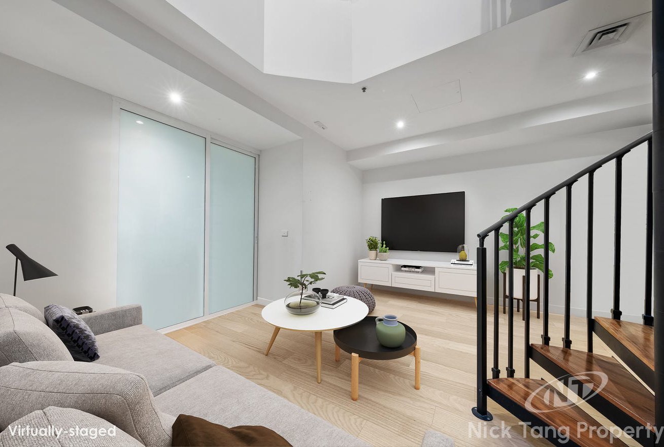 Modern living room with white walls, light wood flooring, a beige sofa with pillows, two round coffee tables, a flat-screen TV on a white stand, green plants, and a staircase with black railing.