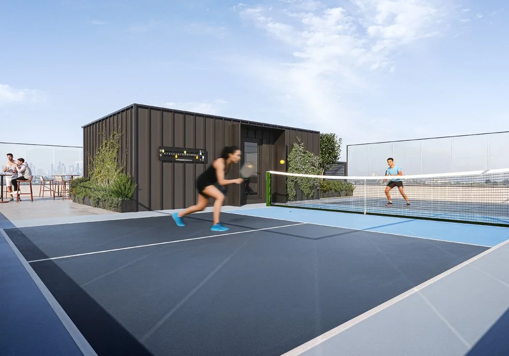 Two people playing tennis on a rooftop court with a city skyline in the background, while two others sit at a table nearby.