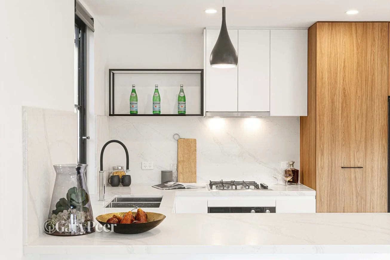 Modern kitchen with white cabinetry, marble backsplash, wooden panel on the right, and a black pendant light hanging above the countertop. On the counter, there is a glass jar with fruit and a bowl with snacks, a cutting board, and some bottles. Thre