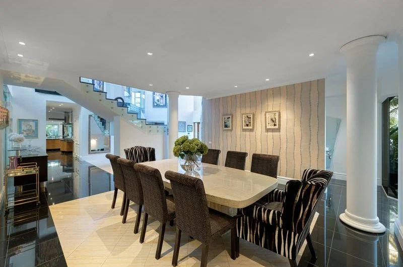 Modern dining room with a large rectangular table, ten chairs, and a floral centerpiece. The space has a staircase, wall art, and large windows.