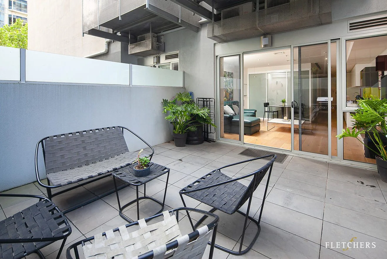 An outdoor balcony with a black metal chairs and small table, potted plants, and sliding glass doors leading to an indoor living room with a couch, coffee table, and dining area.