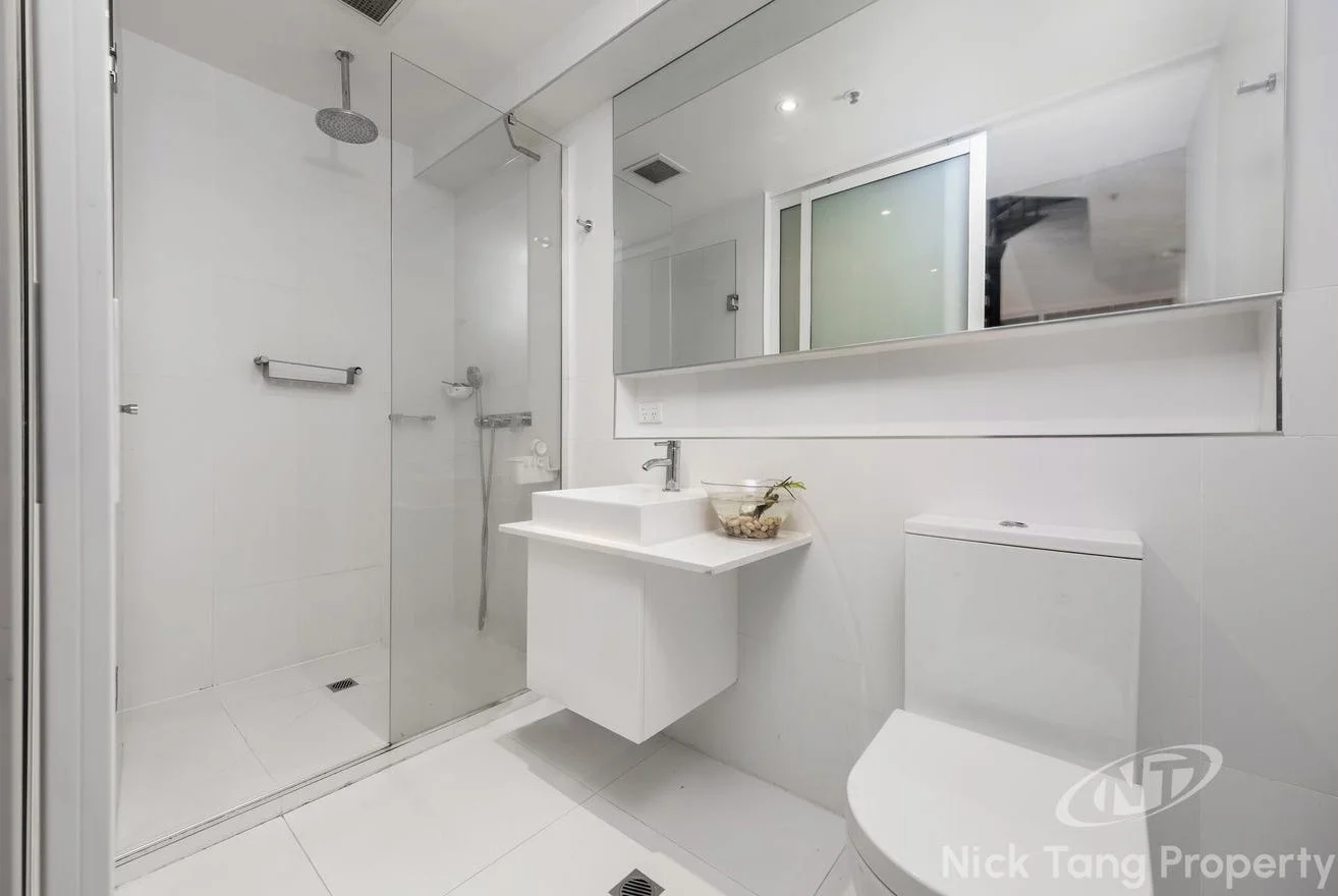 A modern, minimalist bathroom featuring a walk-in shower with glass doors, a wall-mounted sink with a decorative bowl, a toilet, and a large mirror. The space is all white with bright lighting.
