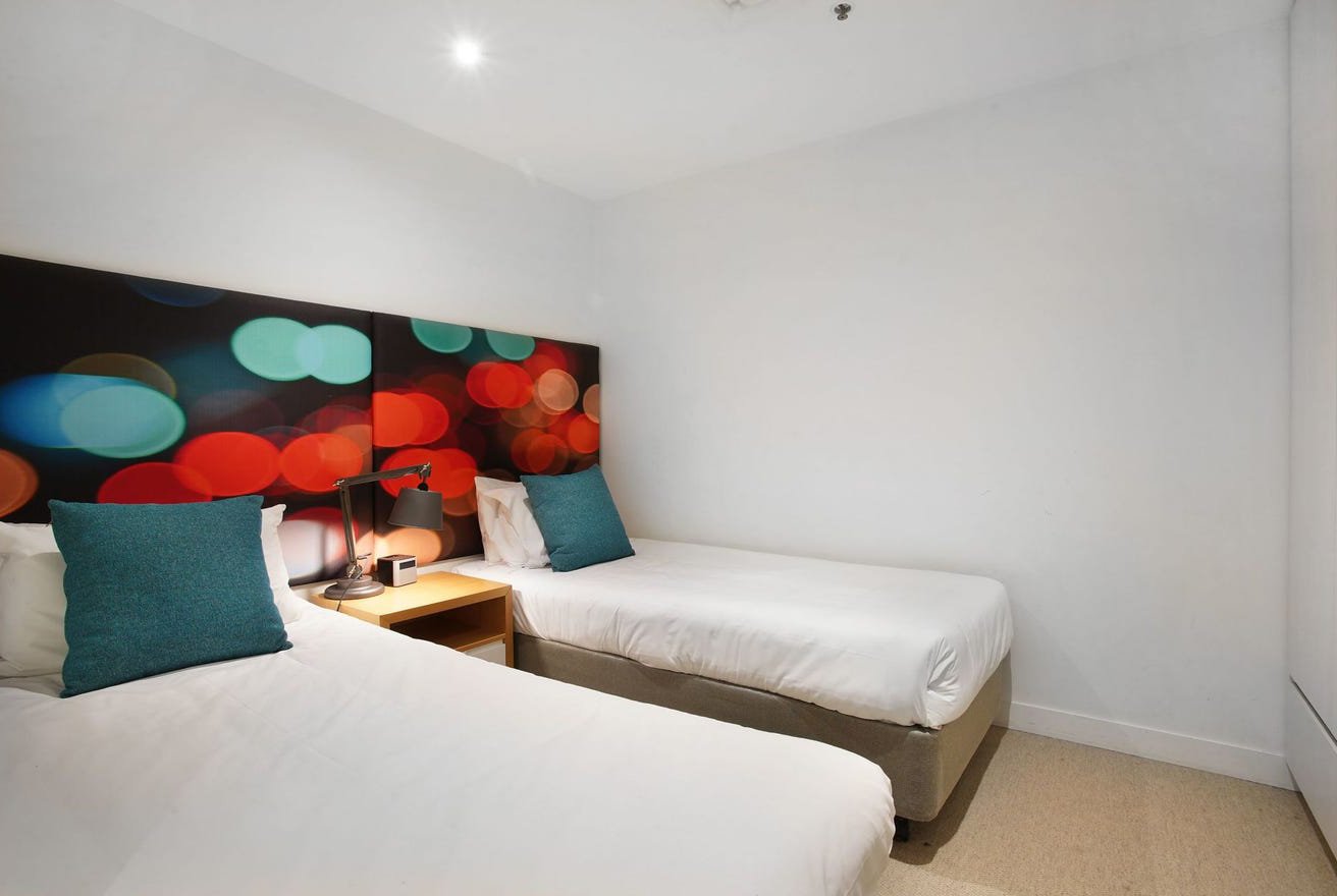 A hotel room with two beds, a nightstand, a lamp, and a headboard with colorful, blurry light pattern.